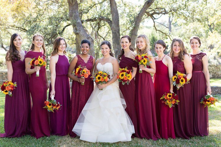 Bridesmaids in burgundy dresses surround a bride in a white gown holding a bouquet outdoors under trees