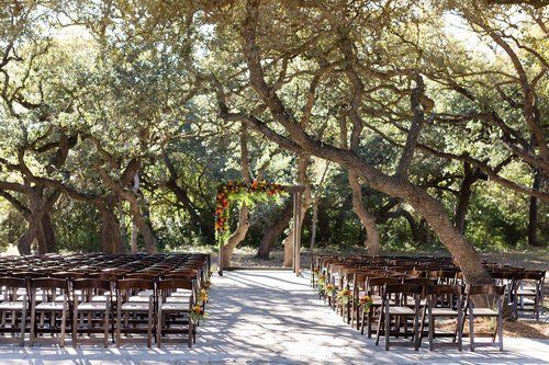 Outdoor wedding ceremony setup beneath oak trees with rows of chairs and a floral arch on a stone path