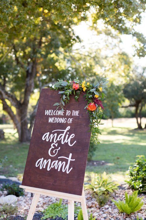 Welcome sign on easel with flowers in a garden, reading “Welcome to the wedding of Angie & Anant”