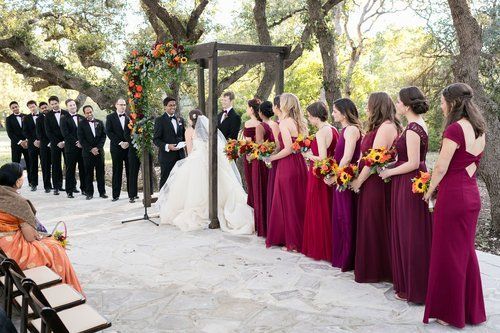 Wedding ceremony under trees with bride and groom facing bridesmaids in burgundy dresses and groomsmen in black suits
