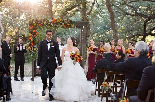 Bride and groom walk down an outdoor wedding aisle with guests seated under trees and string lights
