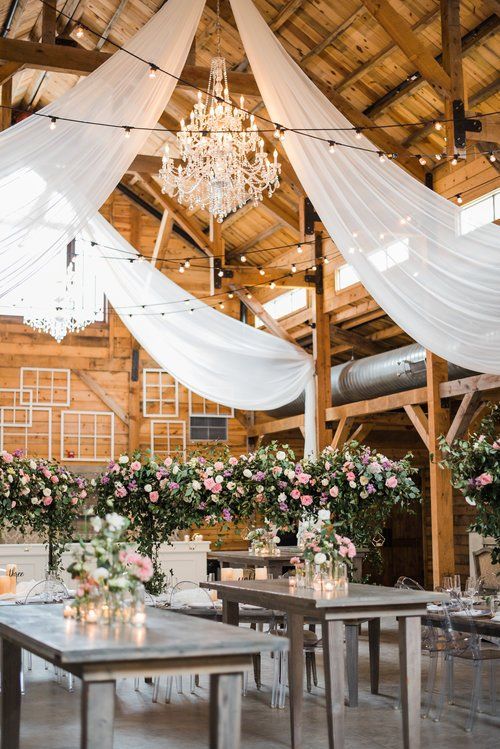 Rustic wedding reception with draped white fabric, chandeliers, flowers, and long tables in a wooden barn