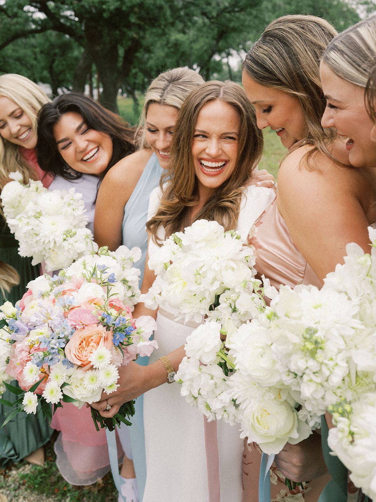 Five smiling women in pastel dresses holding white and pink bouquets outdoors.