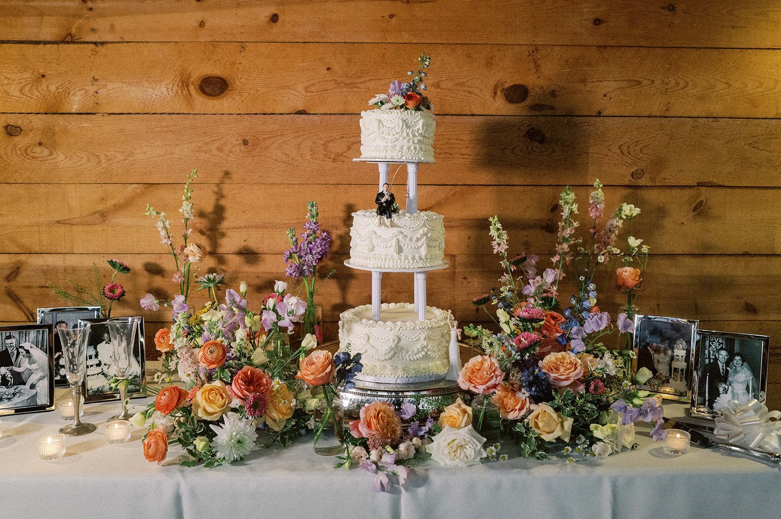 Three-tier white wedding cake with flowers, surrounded by colorful floral arrangements on a rustic wooden backdrop
