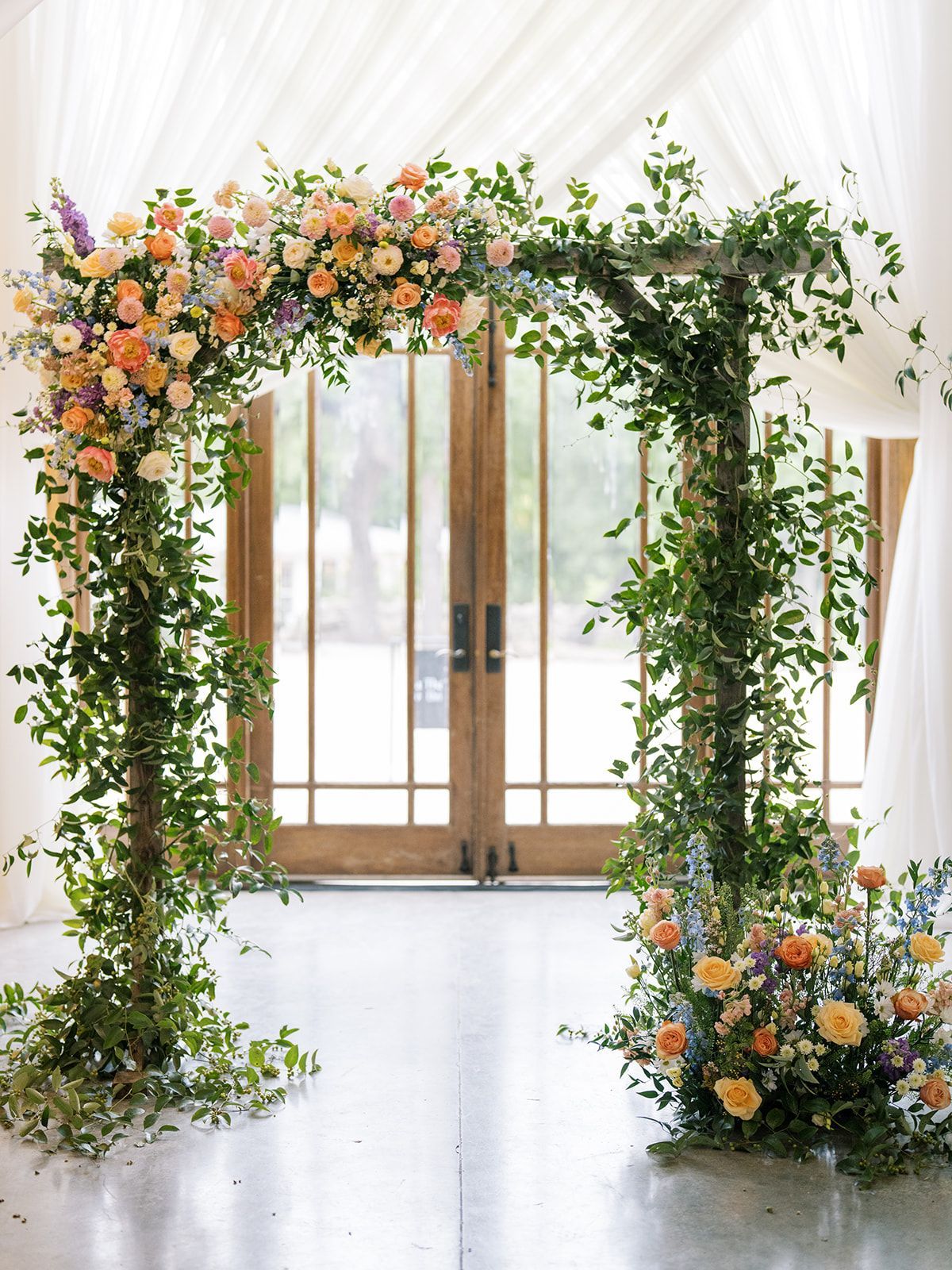 Flower-covered archway framing double wooden doors in a bright indoor wedding venue