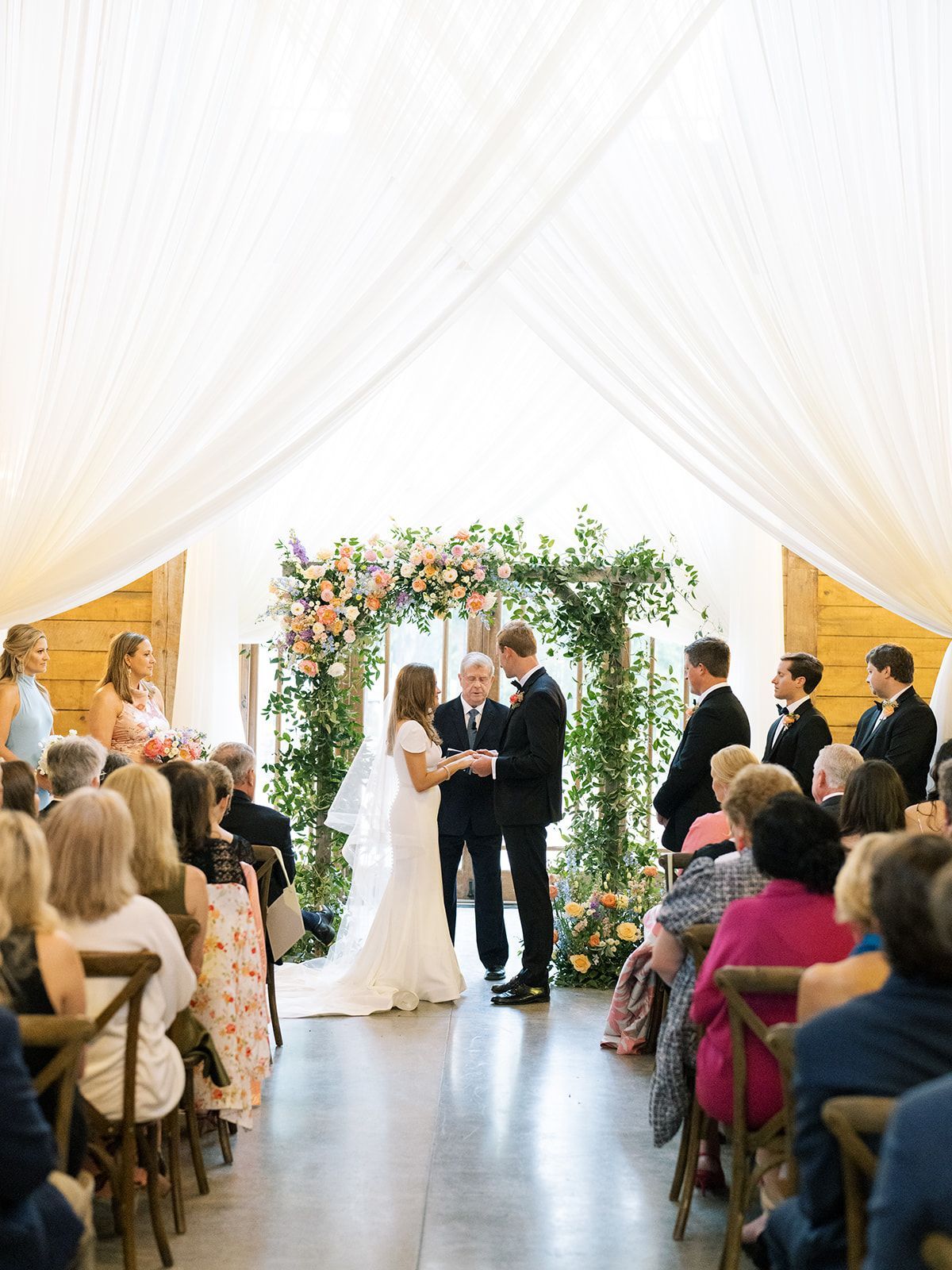 Wedding ceremony under a floral arch, with the couple and officiant at the altar and guests seated on both sides.