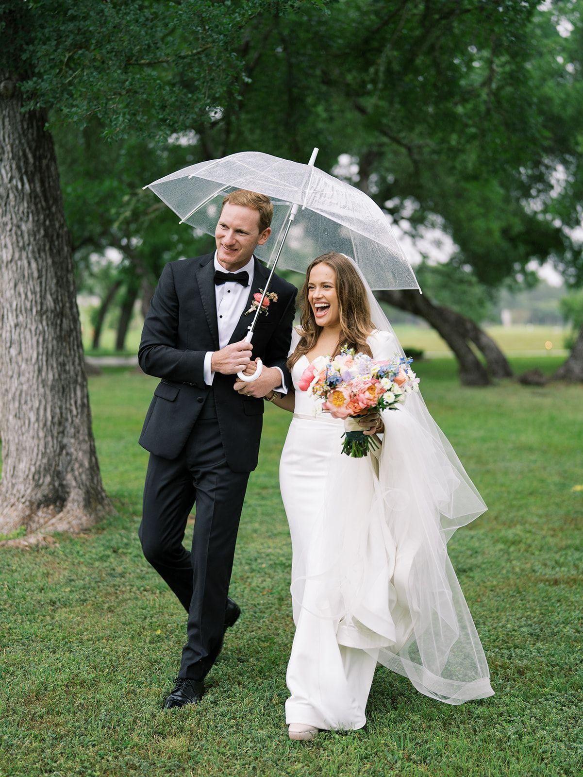 Bride and groom walking under a clear umbrella in a rainy park, both smiling and dressed for a wedding.