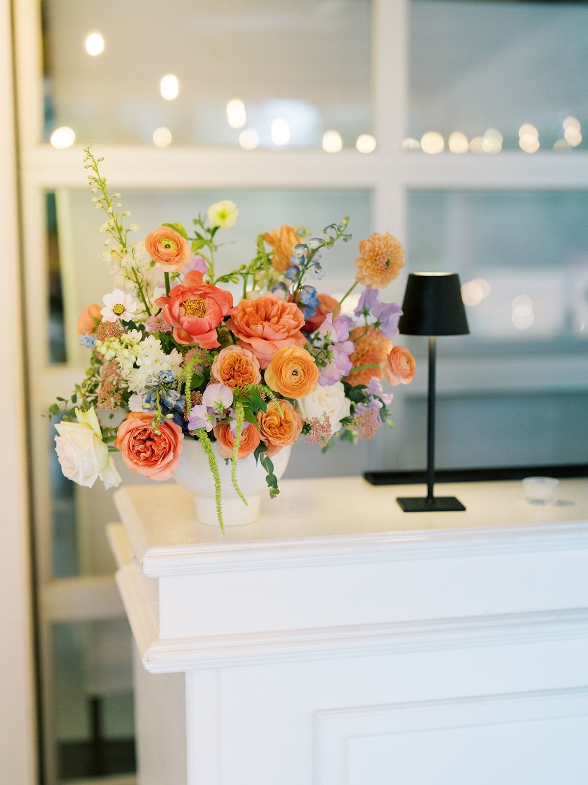 Colorful floral arrangement on a white mantel beside a black lamp in a bright room