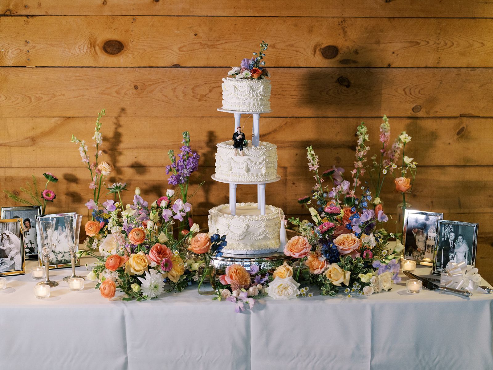 Rustic dessert table with a tall white tiered cake, colorful flowers, and framed photos against a wood wall