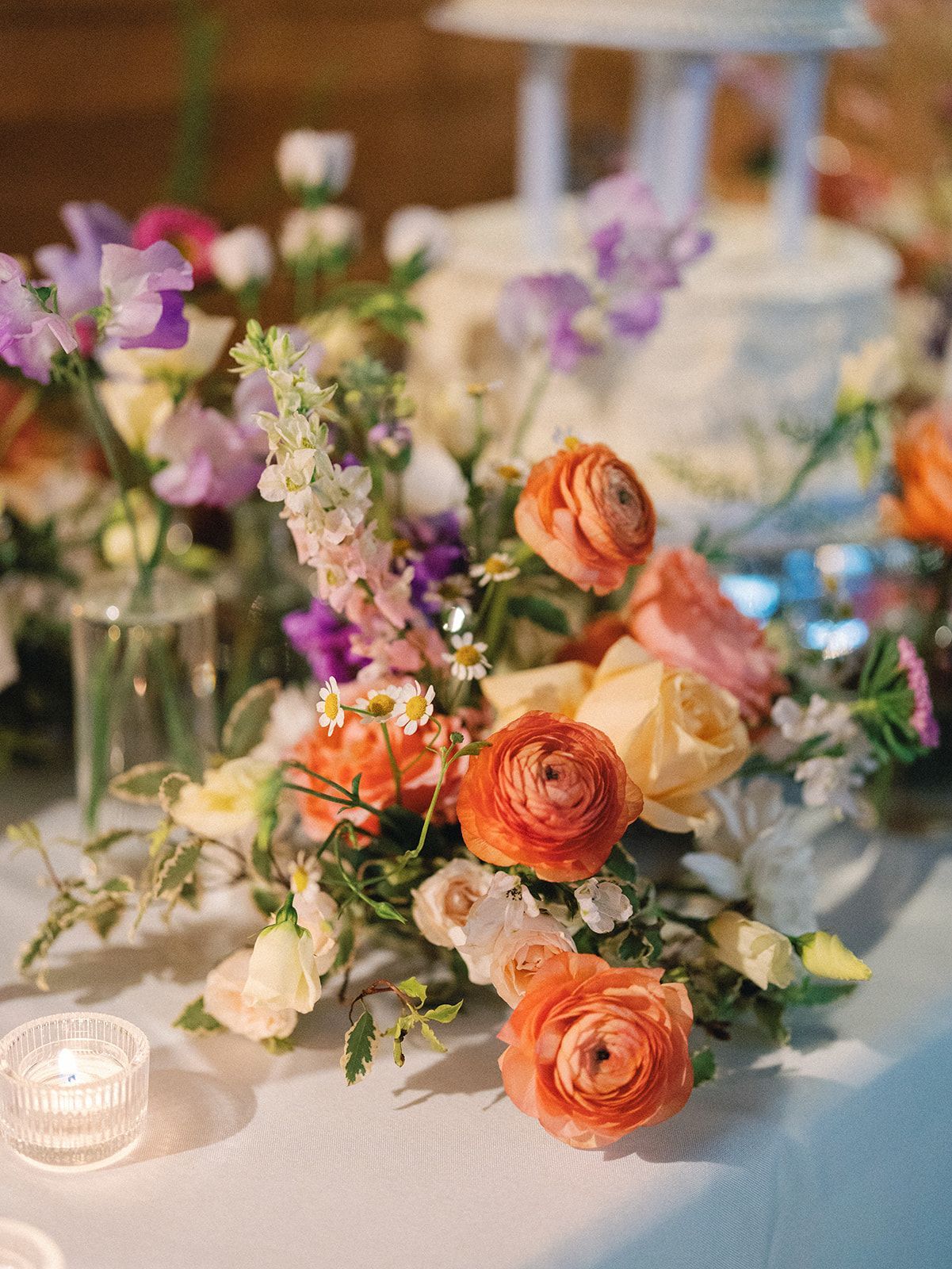 Pastel floral centerpiece with orange roses, purple flowers, and a lit candle on a white table