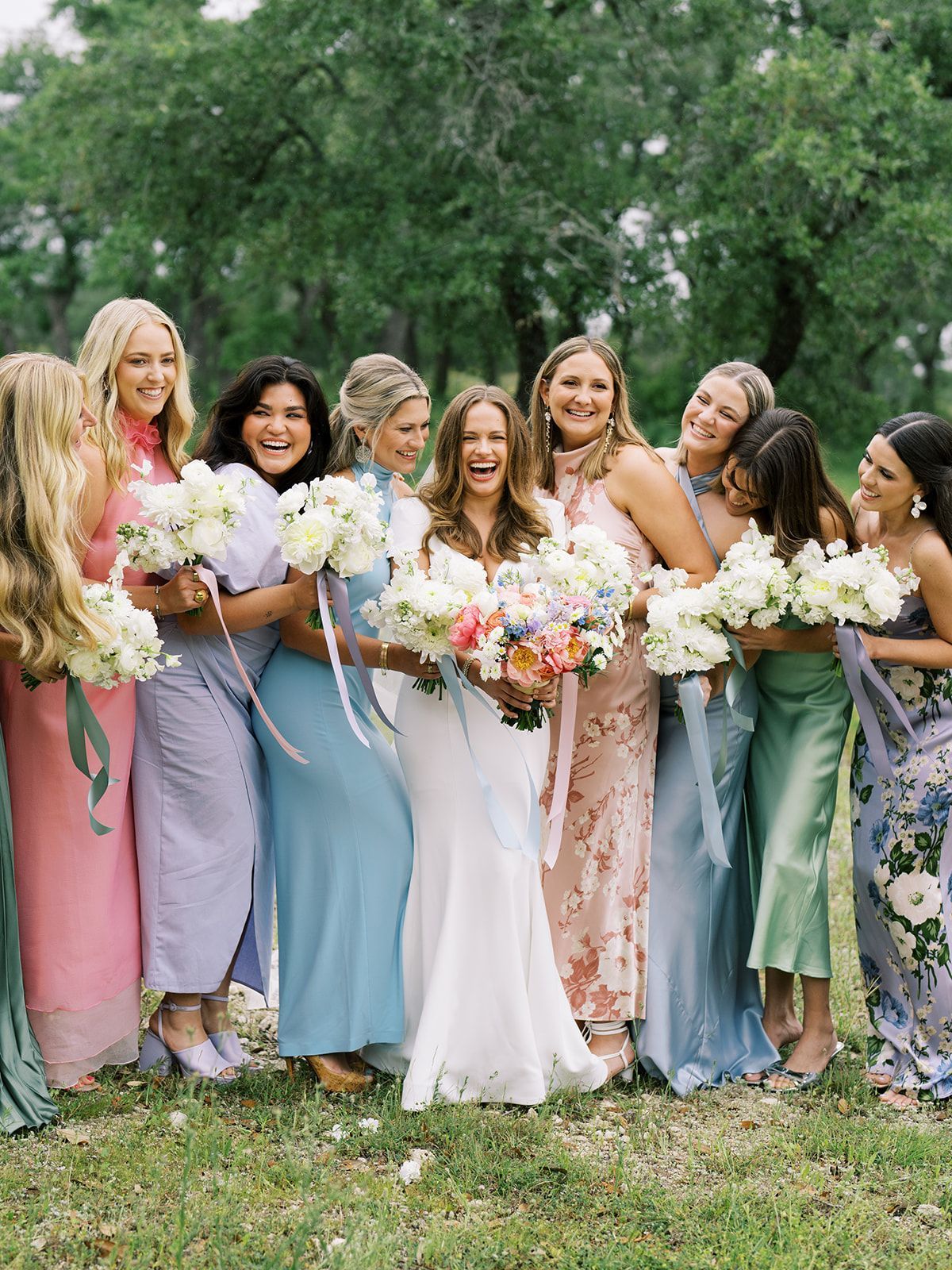 Group of people in pastel dresses holding bouquets outdoors on a grassy lawn, smiling together.