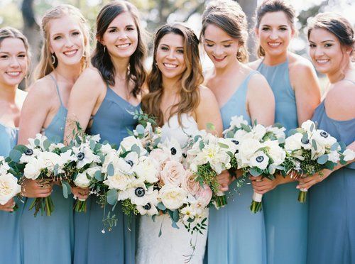 Seven women in blue dresses holding white and blush flower bouquets, smiling outdoors.