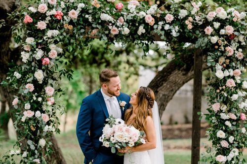 Bride and groom kissing under a floral arch outdoors, holding a pastel bouquet