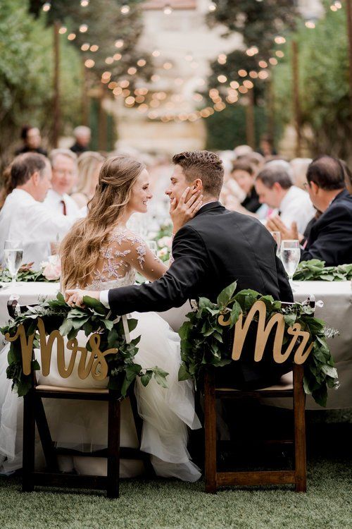 Wedding couple seated at a reception table with greenery, facing guests in an outdoor garden setting