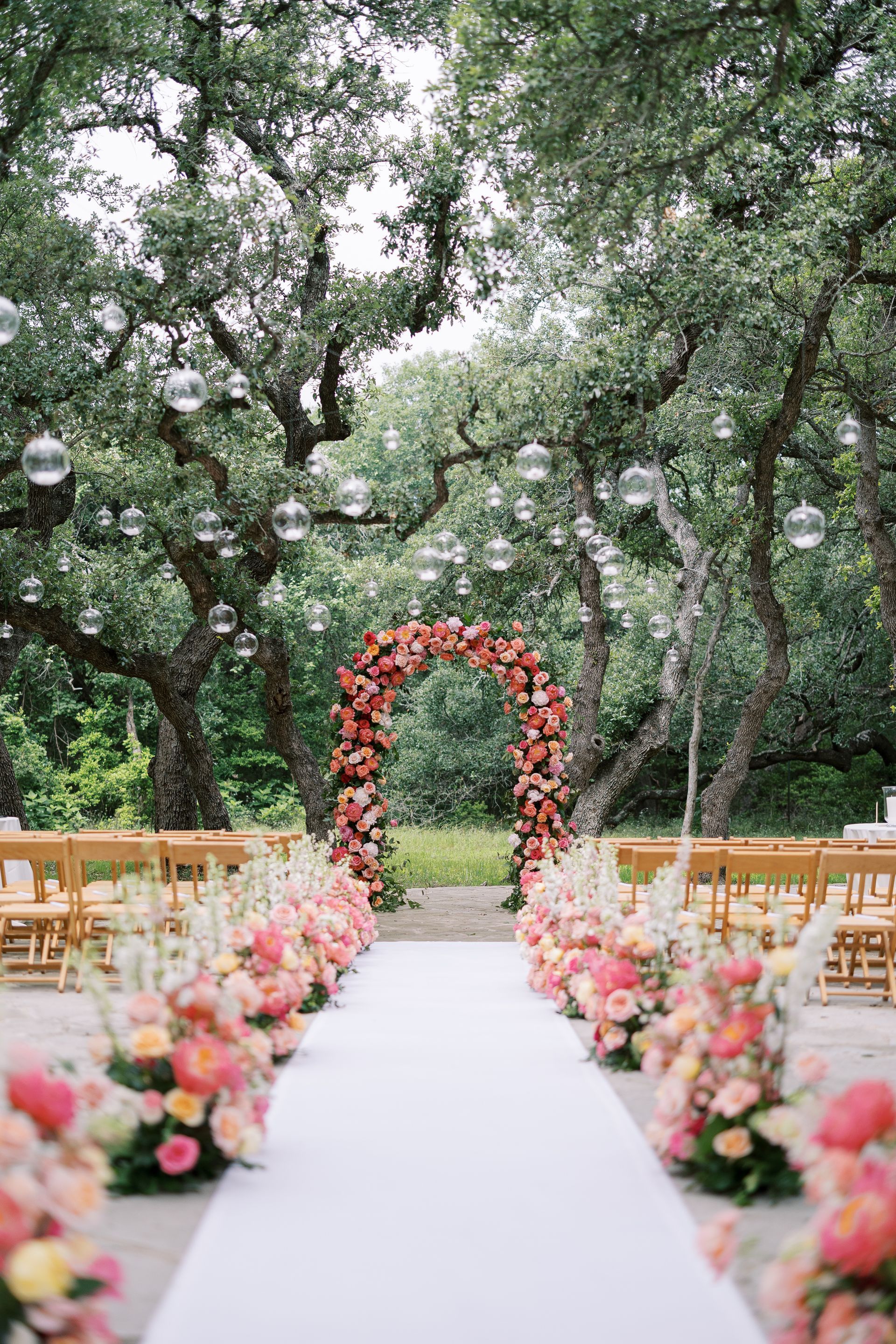 Outdoor wedding aisle with white runner, pink floral arrangements, and a floral arch under trees