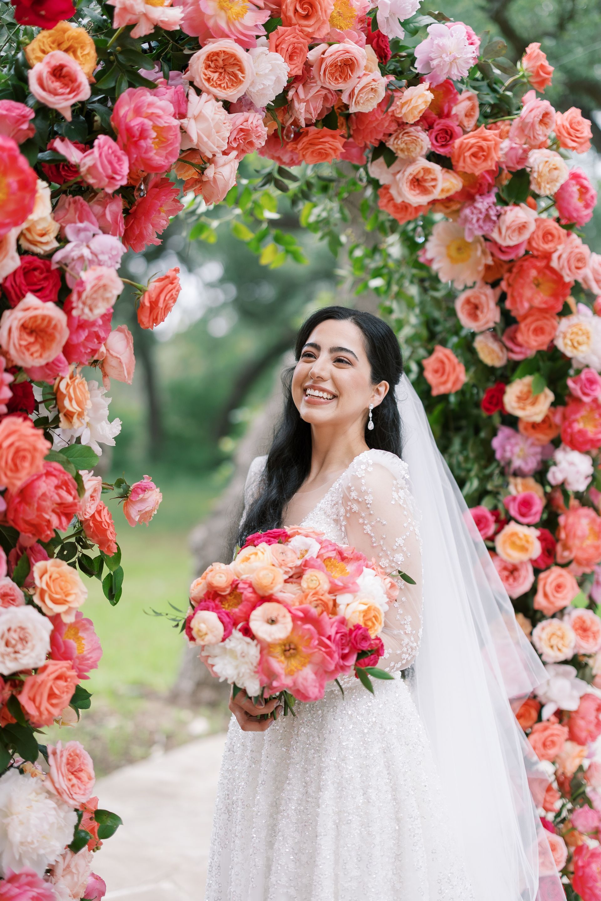 Bride in white gown and veil holding a pink bouquet under a floral arch outdoors