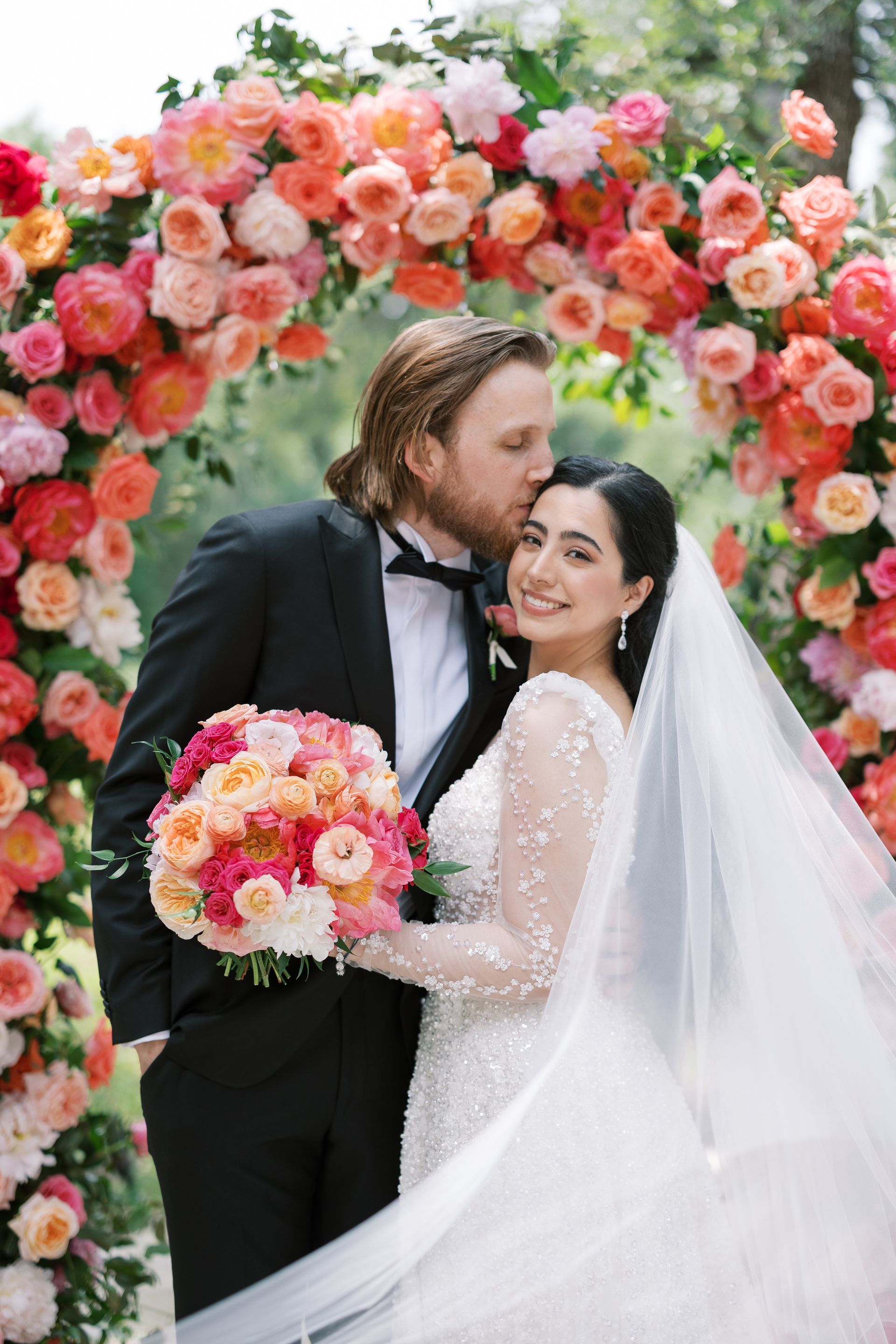 Wedding couple kissing under a floral arch, bride in white veil holding a pink bouquet
