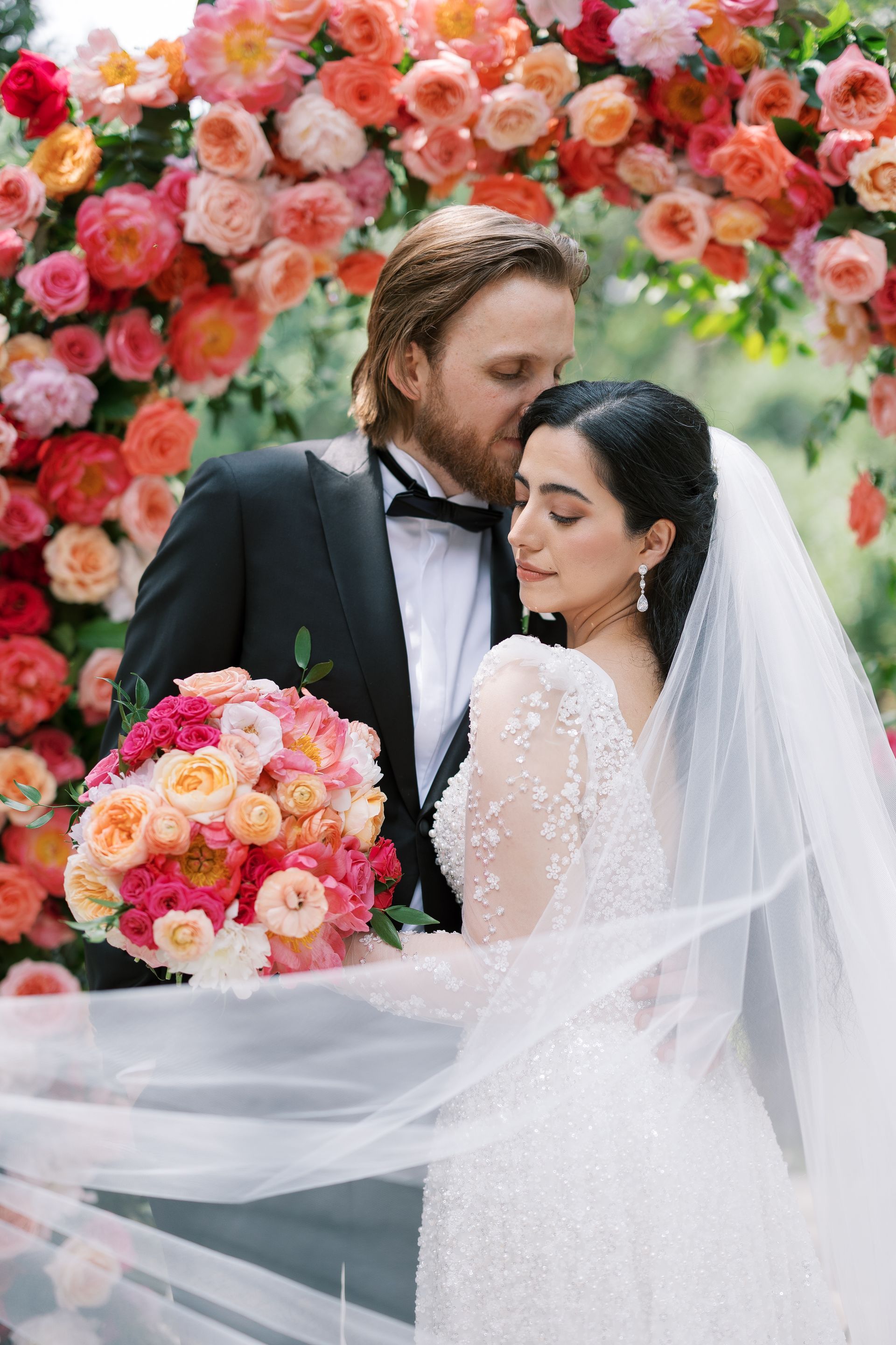 Bride and groom embrace beneath a floral arch, with pink roses and the bride’s white veil flowing softly.