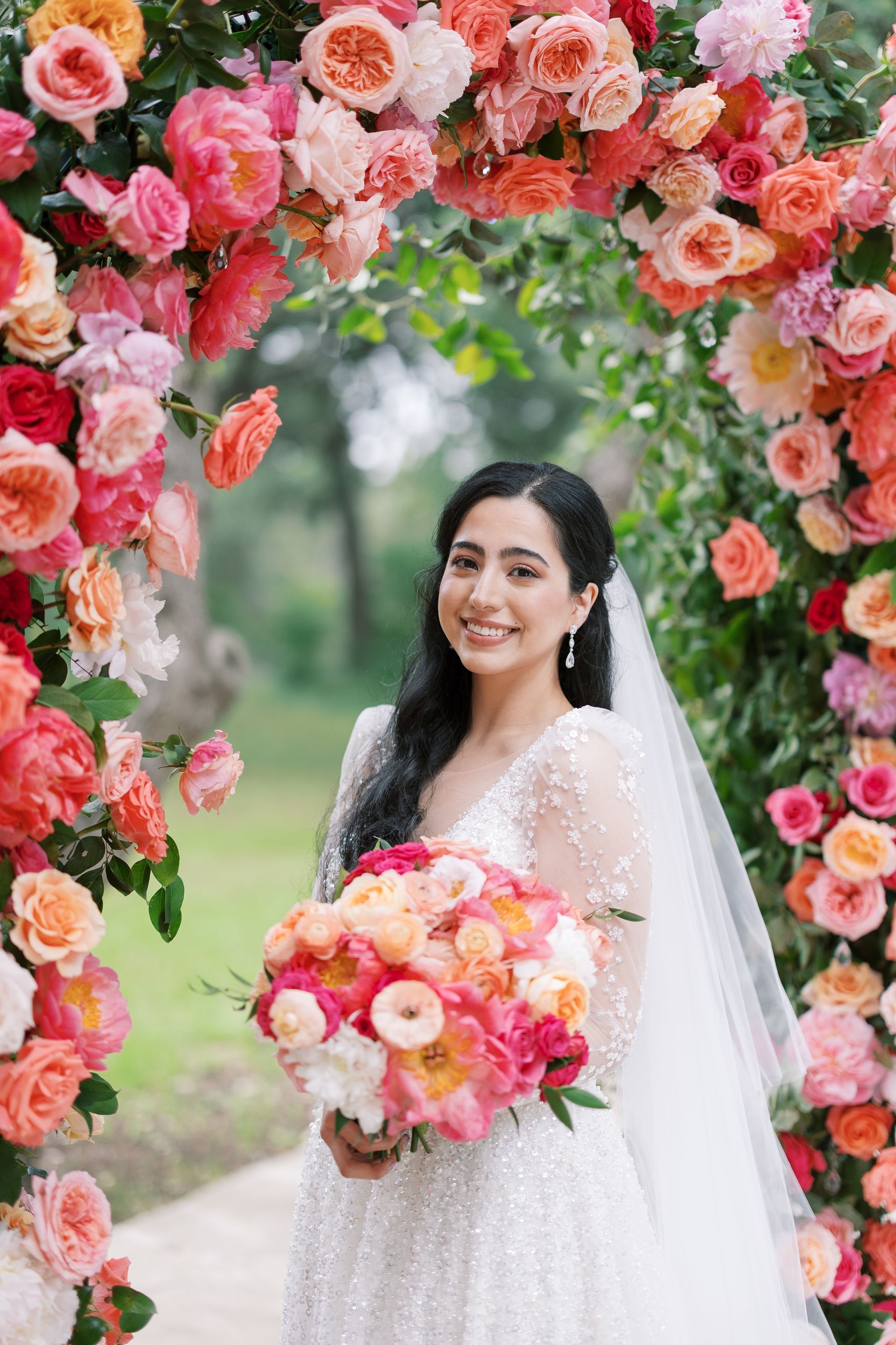 Bride in white dress holding a pink bouquet beneath a floral arch outdoors