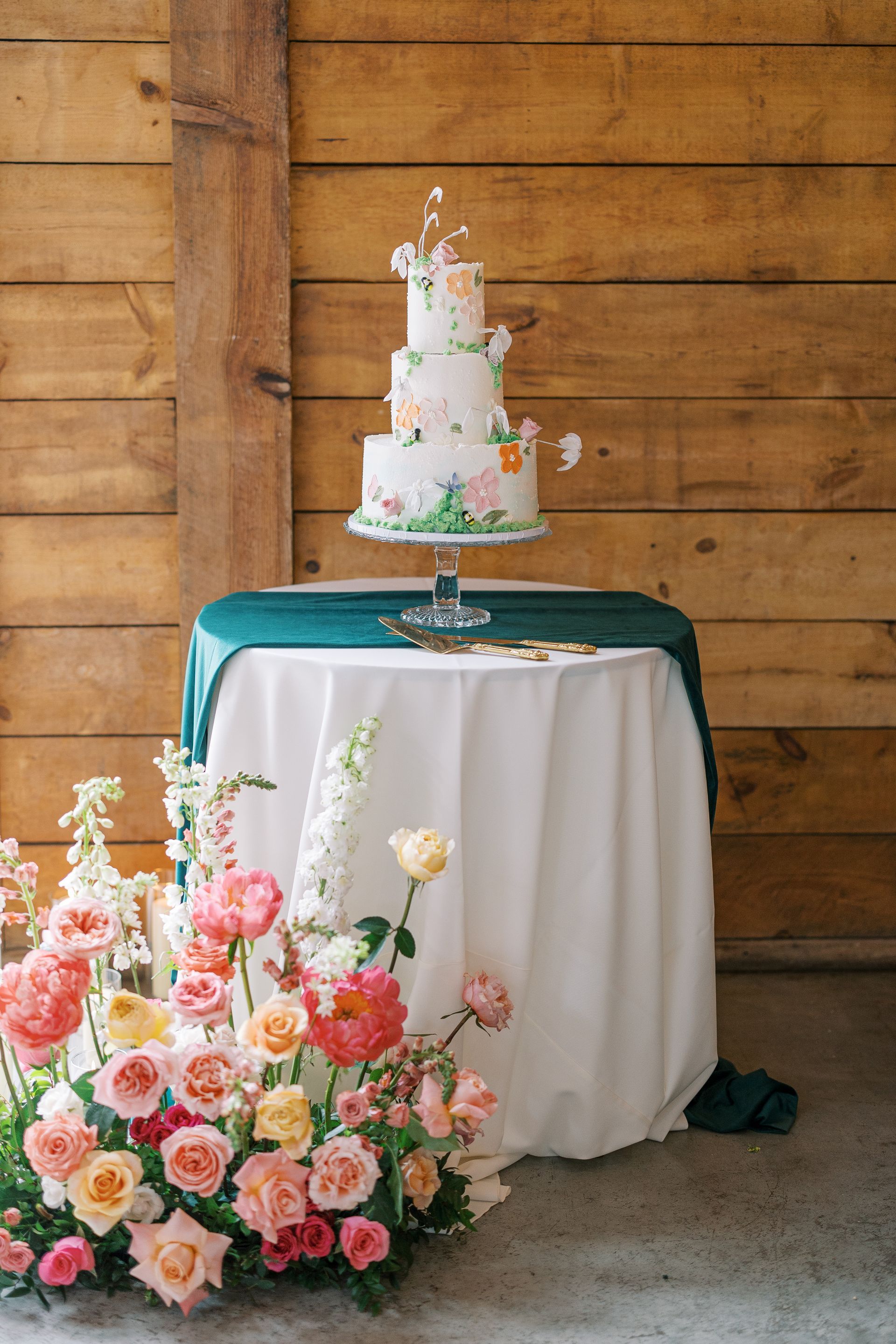 Tall white tiered cake on a green-draped table beside pink and peach flowers at a rustic wooden venue