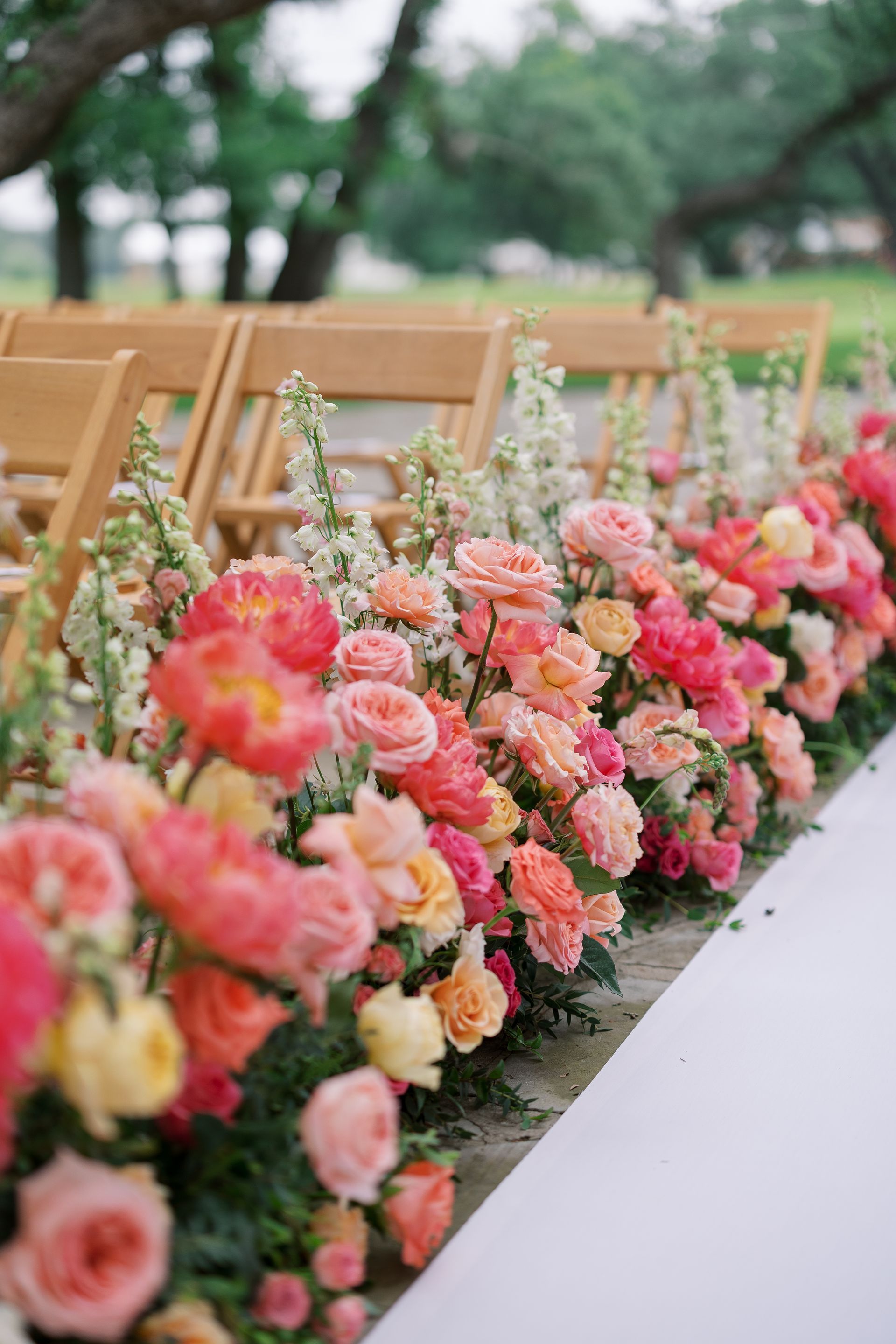 Pink, peach, and cream floral arrangements line wooden chairs along an outdoor aisle.