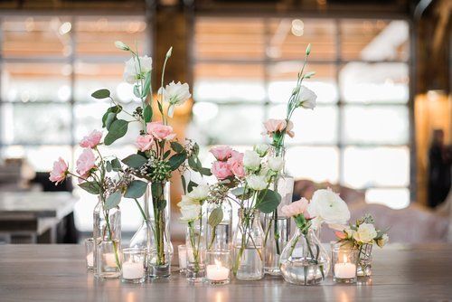 Glass vases of pink and white flowers on a wooden table in a bright, blurred event space