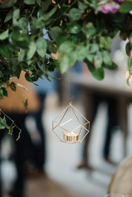Small gold geometric hanging candle holder beneath green leaves, with a blurred indoor background.