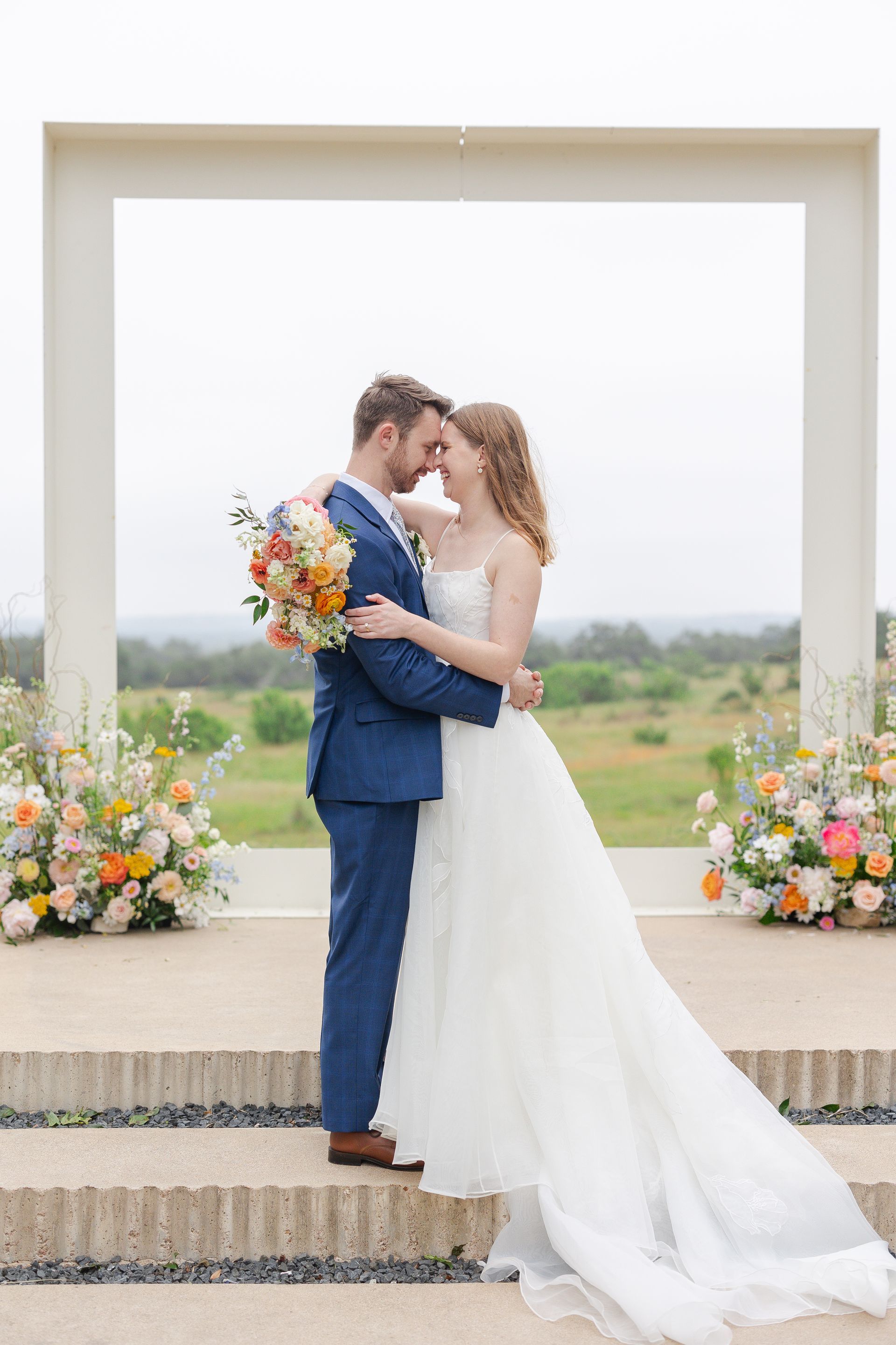 Bride and groom embracing under a white wedding arch with floral arrangements outdoors