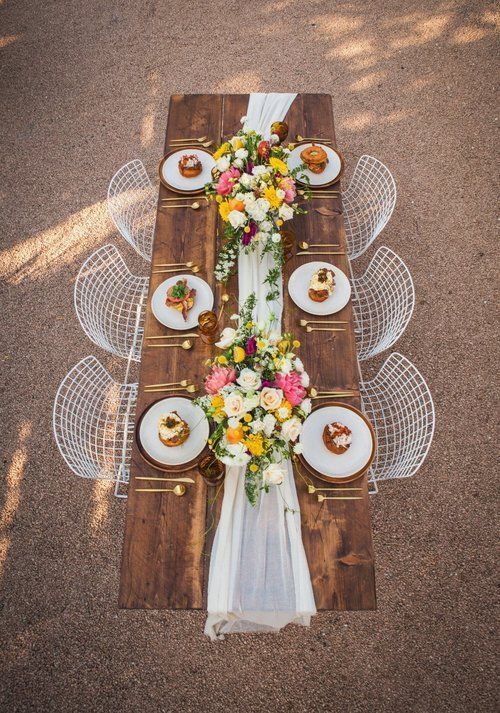 Rustic outdoor table setting with white chairs, floral centerpieces, and place settings on a wooden table runner