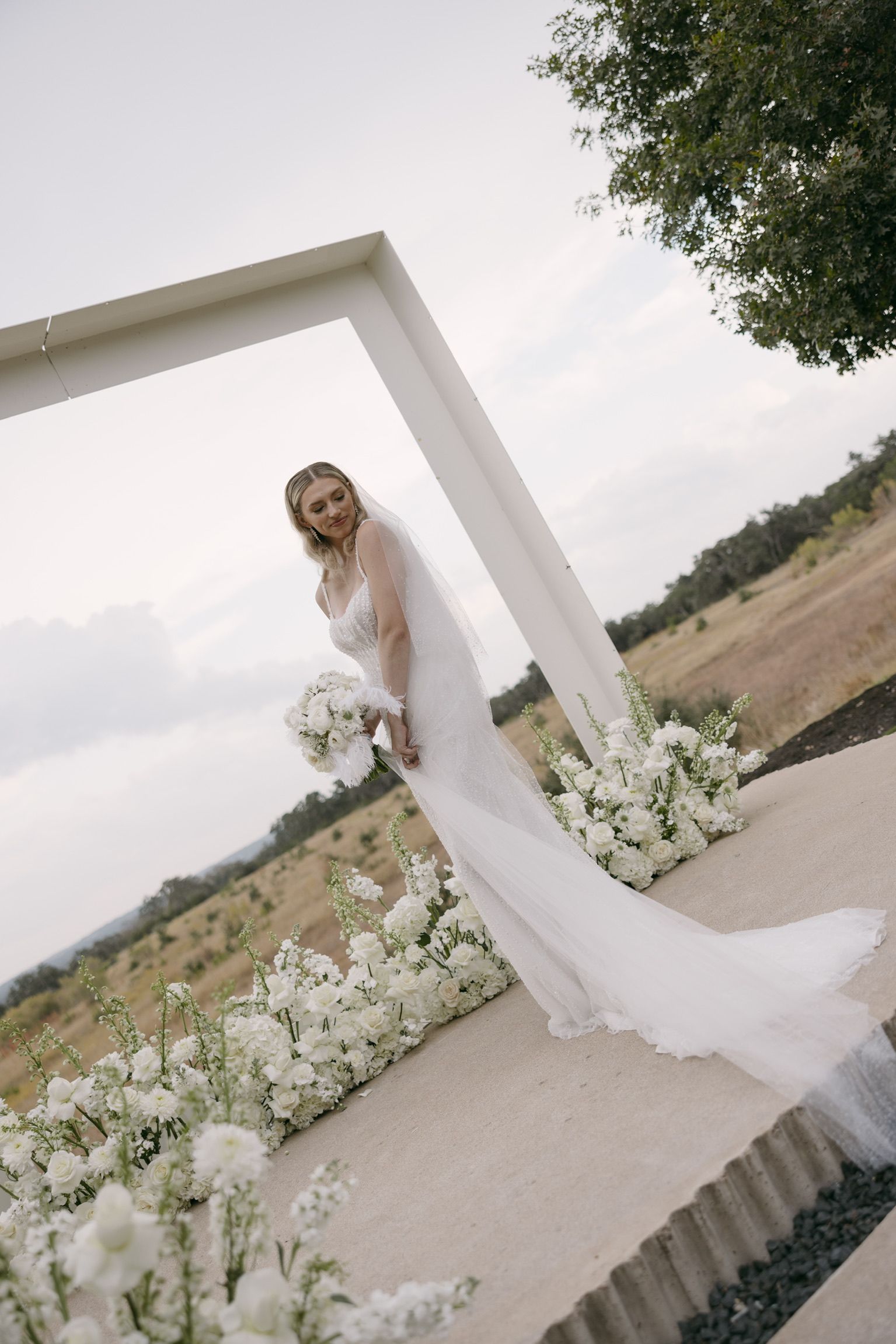 Bride in a white dress holding a bouquet under a floral arch outdoors.