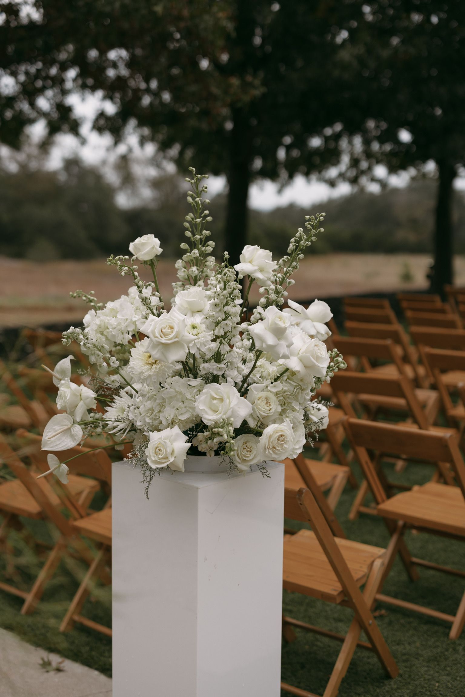 White floral arrangement on a pedestal among rows of wooden chairs outdoors