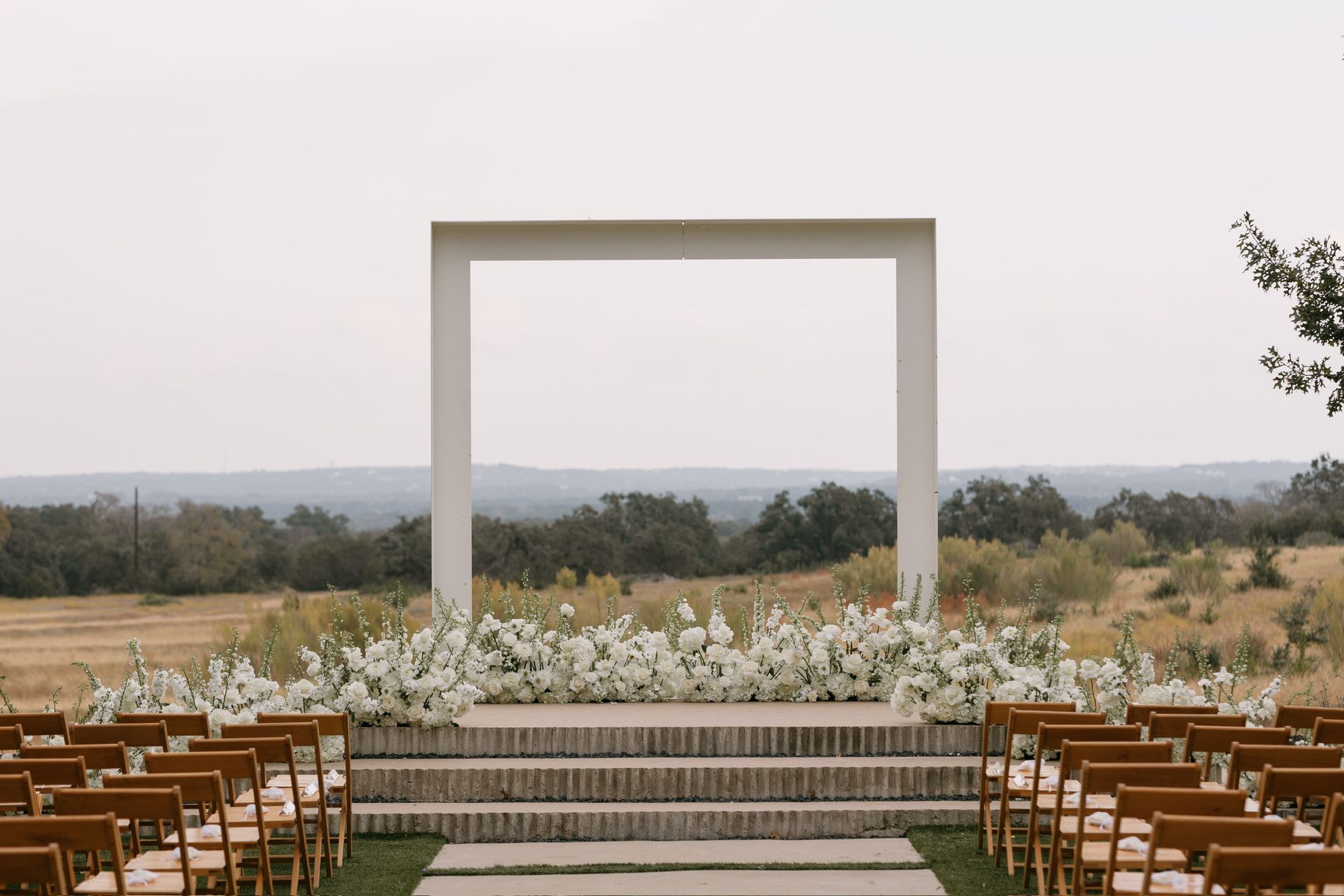 Outdoor wedding ceremony setup with white arch, floral aisle, and rows of wooden chairs overlooking a landscape