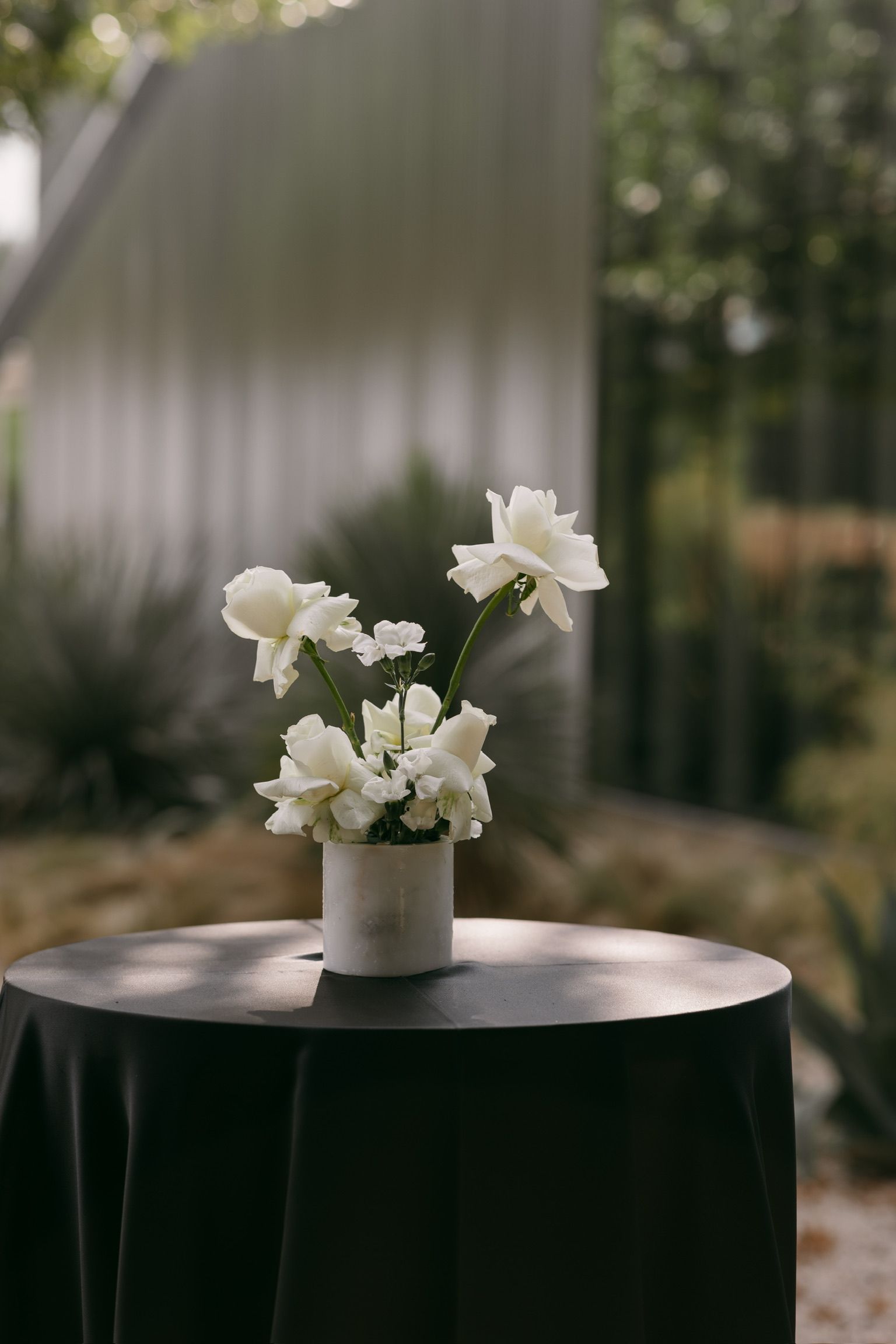White flowers in a small vase on a round black table, with a blurred garden backdrop