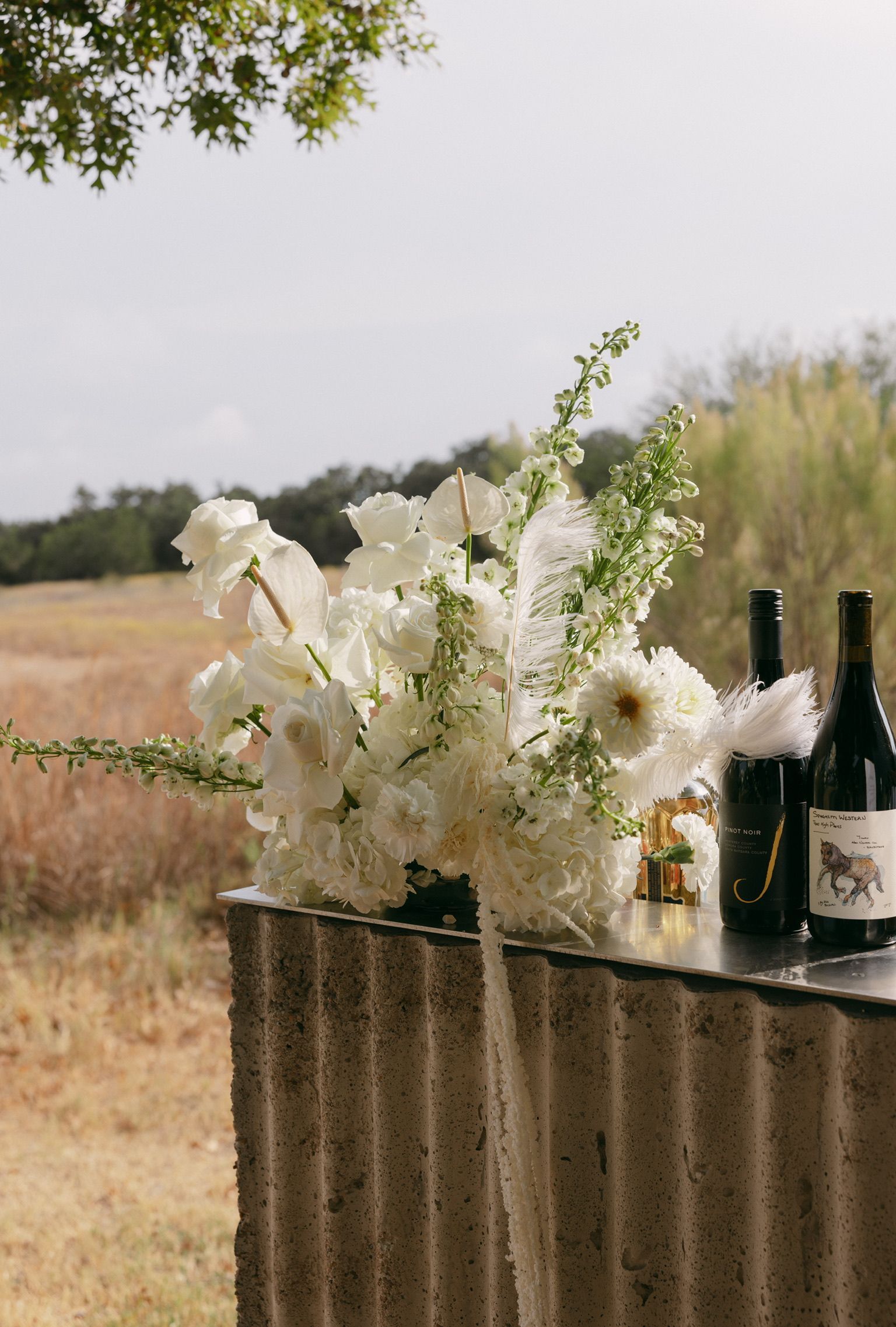 White flowers and wine bottles on a rustic table outdoors in a field.