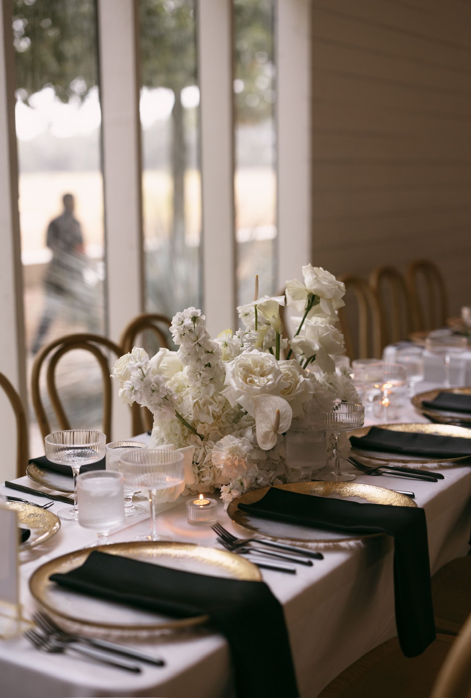 Elegant dining table set with white floral centerpiece, black napkins, and gold chairs by a window