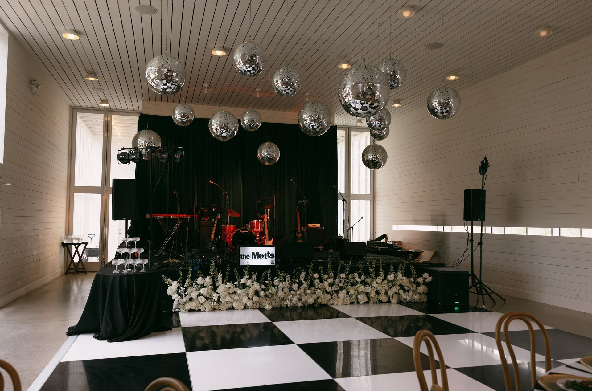 Indoor event hall with black-and-white checkered floor, stage, hanging disco balls, and tables decorated with flowers.