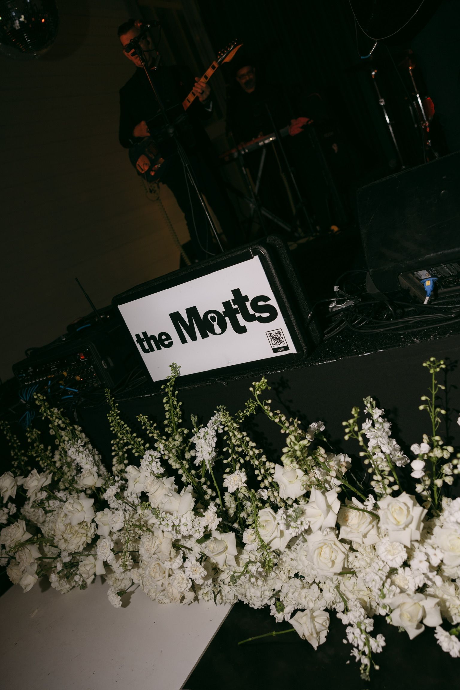 The Mots sign behind a white flower arrangement on a dark stage