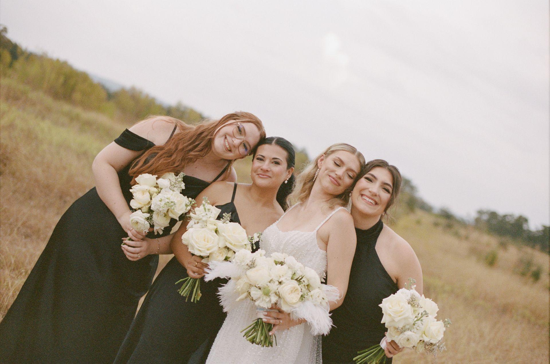 Four women in dresses holding bouquets, smiling in a grassy field outdoors.