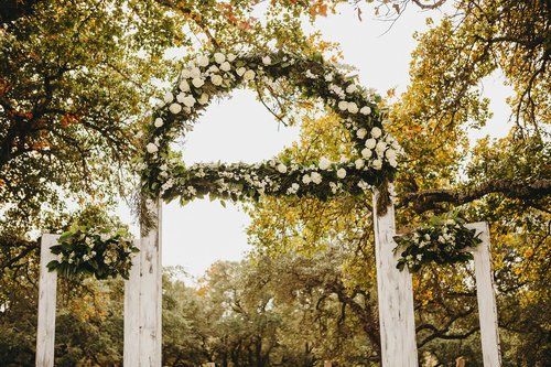 White floral archway outdoors among trees in a sunlit garden setting