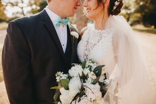 Bride and groom smiling outdoors, holding a white bouquet in soft sunlight