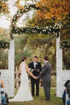 Wedding couple and officiant under a floral arch outdoors, holding hands during the ceremony.