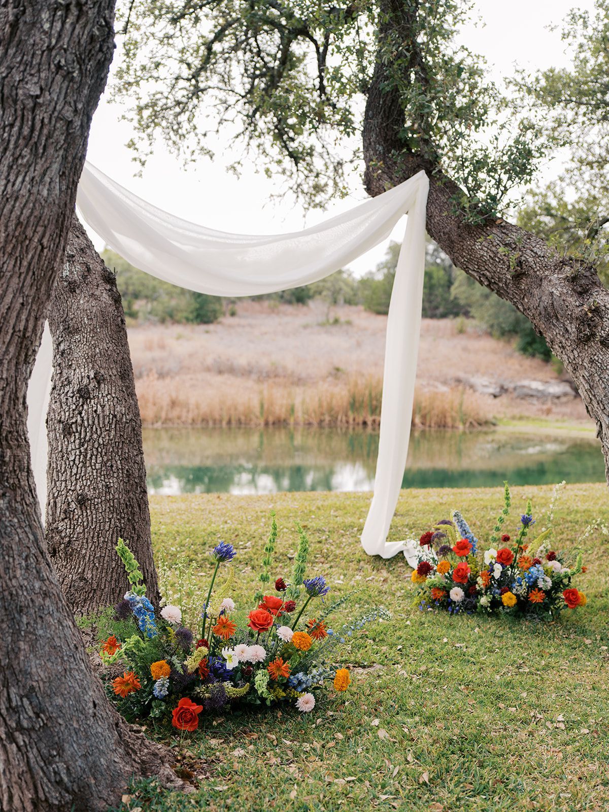 White drape between trees over a lakeside lawn with colorful flower arrangements.