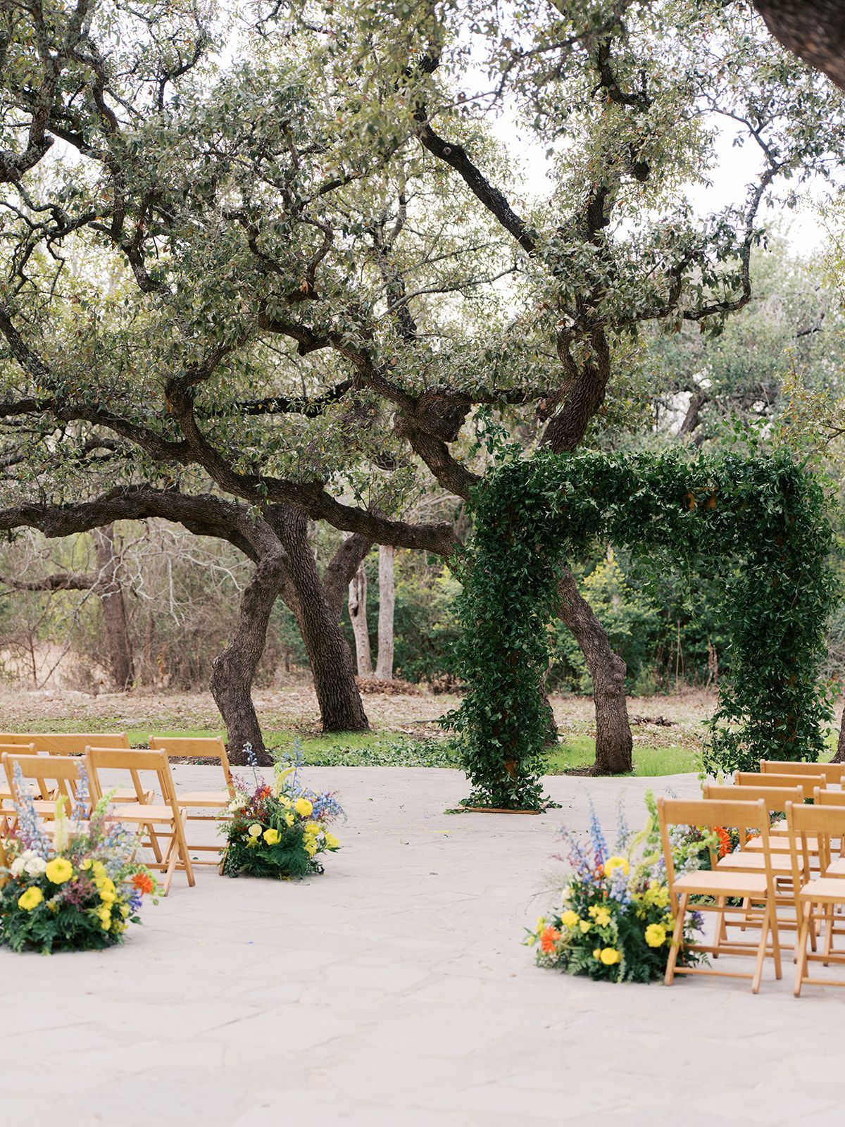 Outdoor wedding ceremony setup with wooden chairs, floral arrangements, and a green arch under large trees