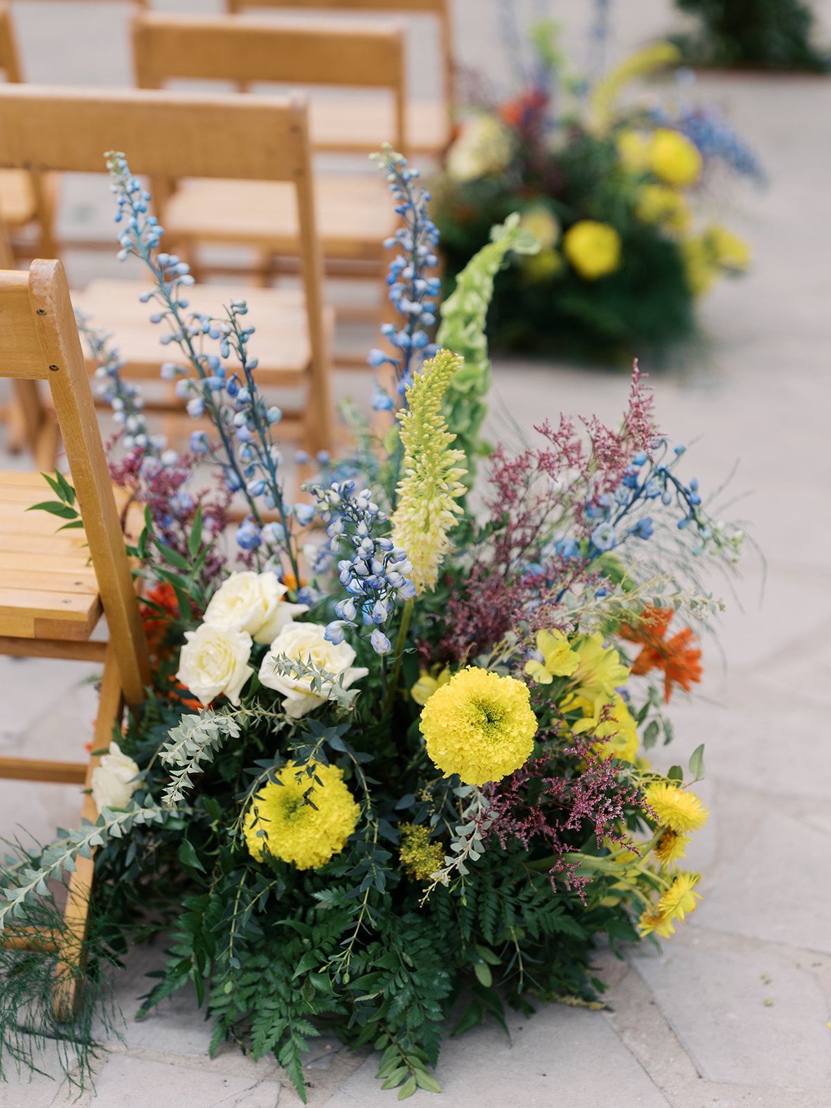 Floral arrangement with yellow, white, blue, and pink flowers beside wooden chairs outdoors
