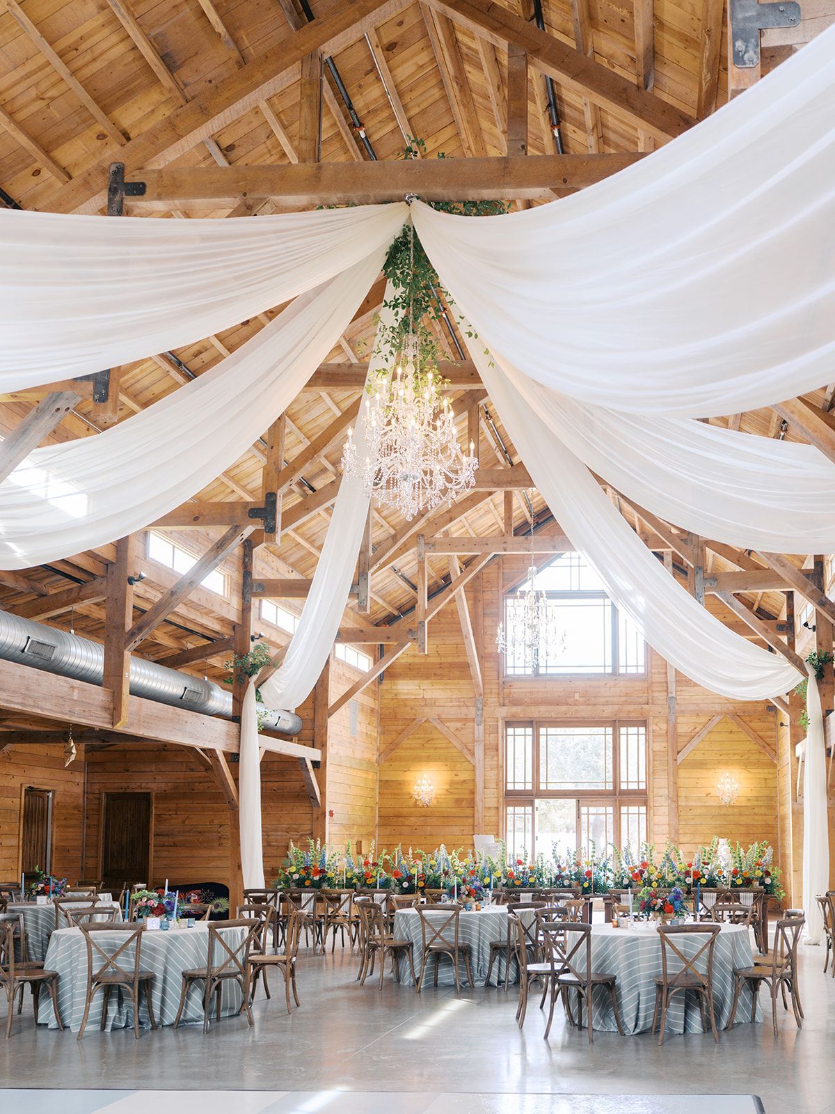Rustic barn wedding reception with white drapes, chandeliers, long tables, and floral centerpieces