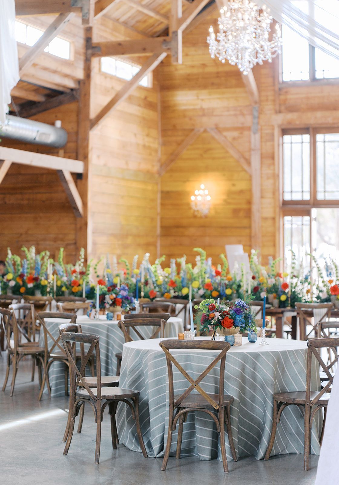 Rustic event hall with round tables, wooden chairs, floral centerpieces, and large windows