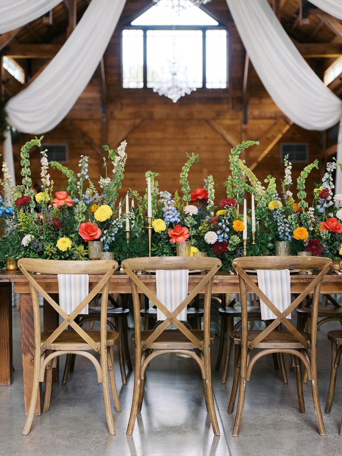 Rustic dining table with colorful floral centerpiece, wood chairs, and white draped fabric backdrop