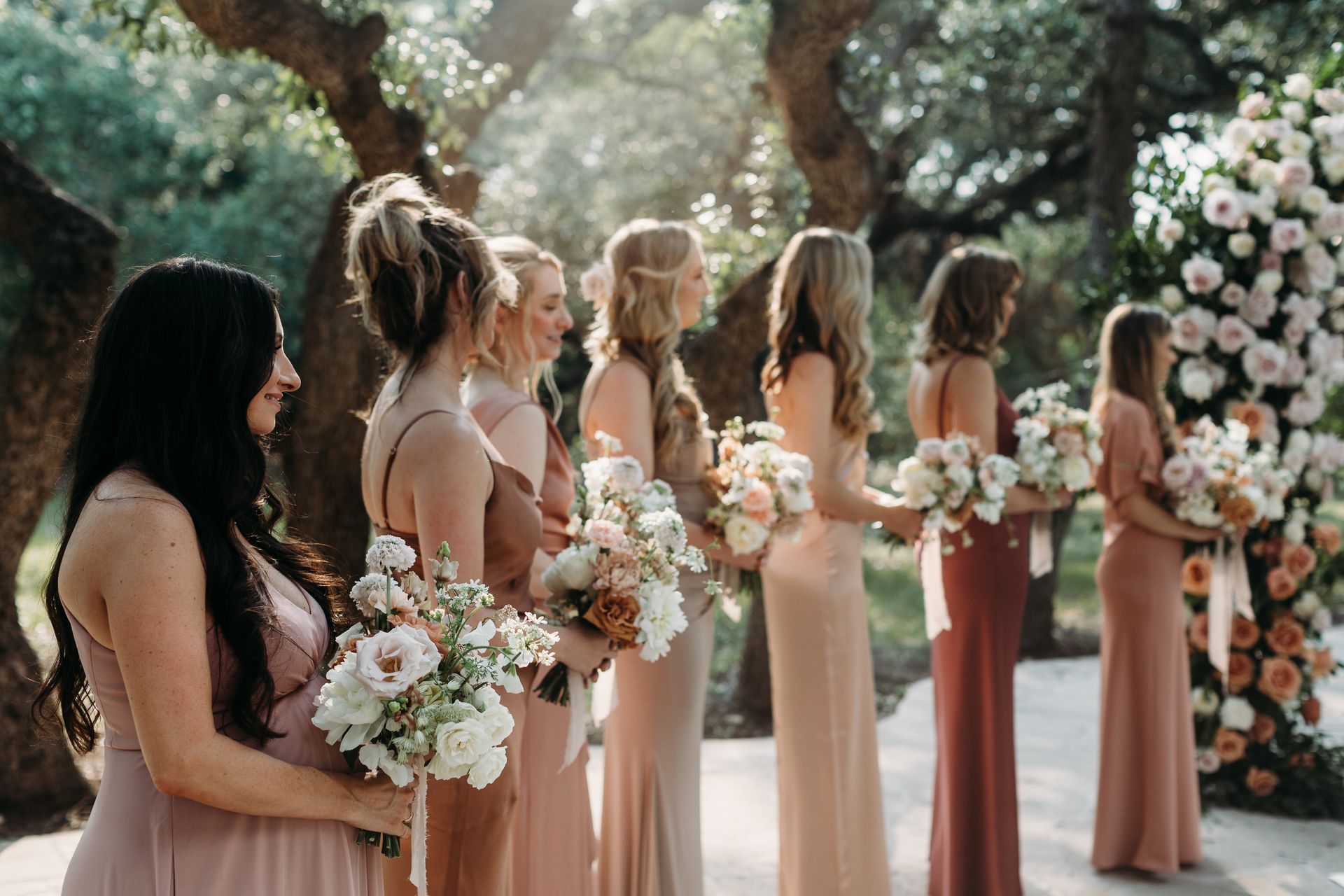 Bridesmaids in blush dresses hold white bouquets at an outdoor wedding ceremony