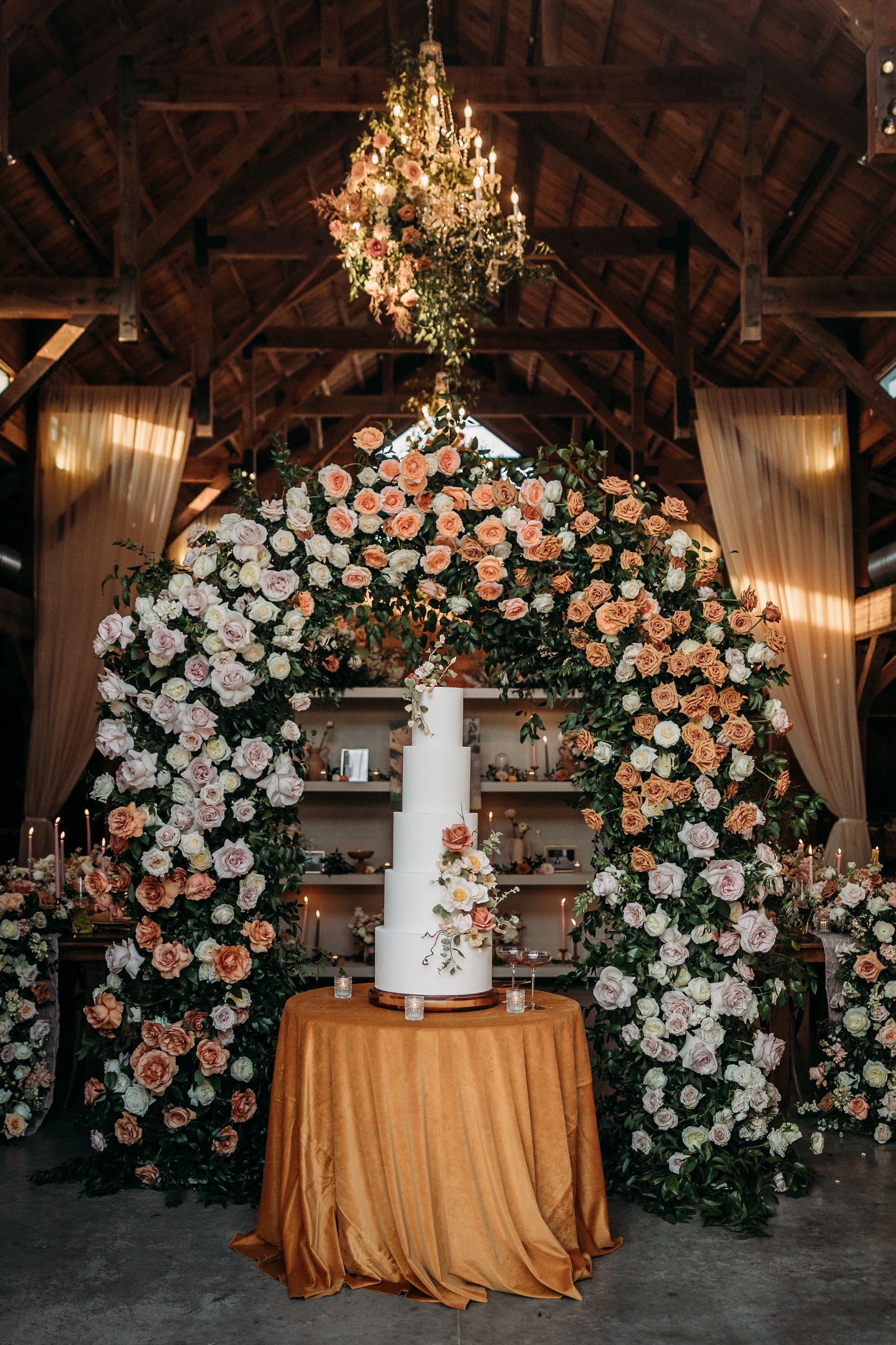 Floral wedding arch with a white tiered cake on a gold-draped table in a rustic barn venue