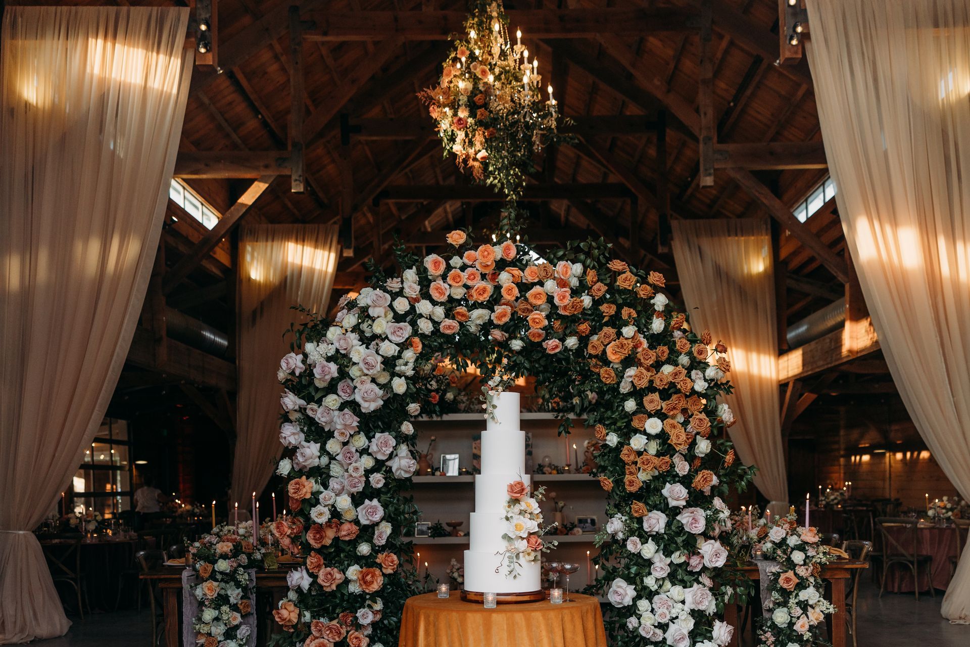 Floral wedding arch with white candles under a rustic barn ceiling, draped curtains, and hanging greenery chandelier